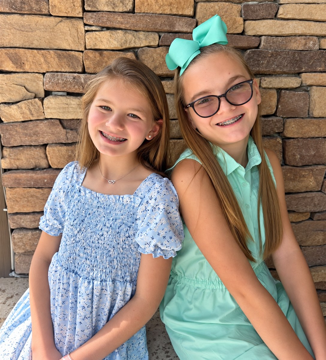 Two smiling sisters seated together in front of a stone wall
